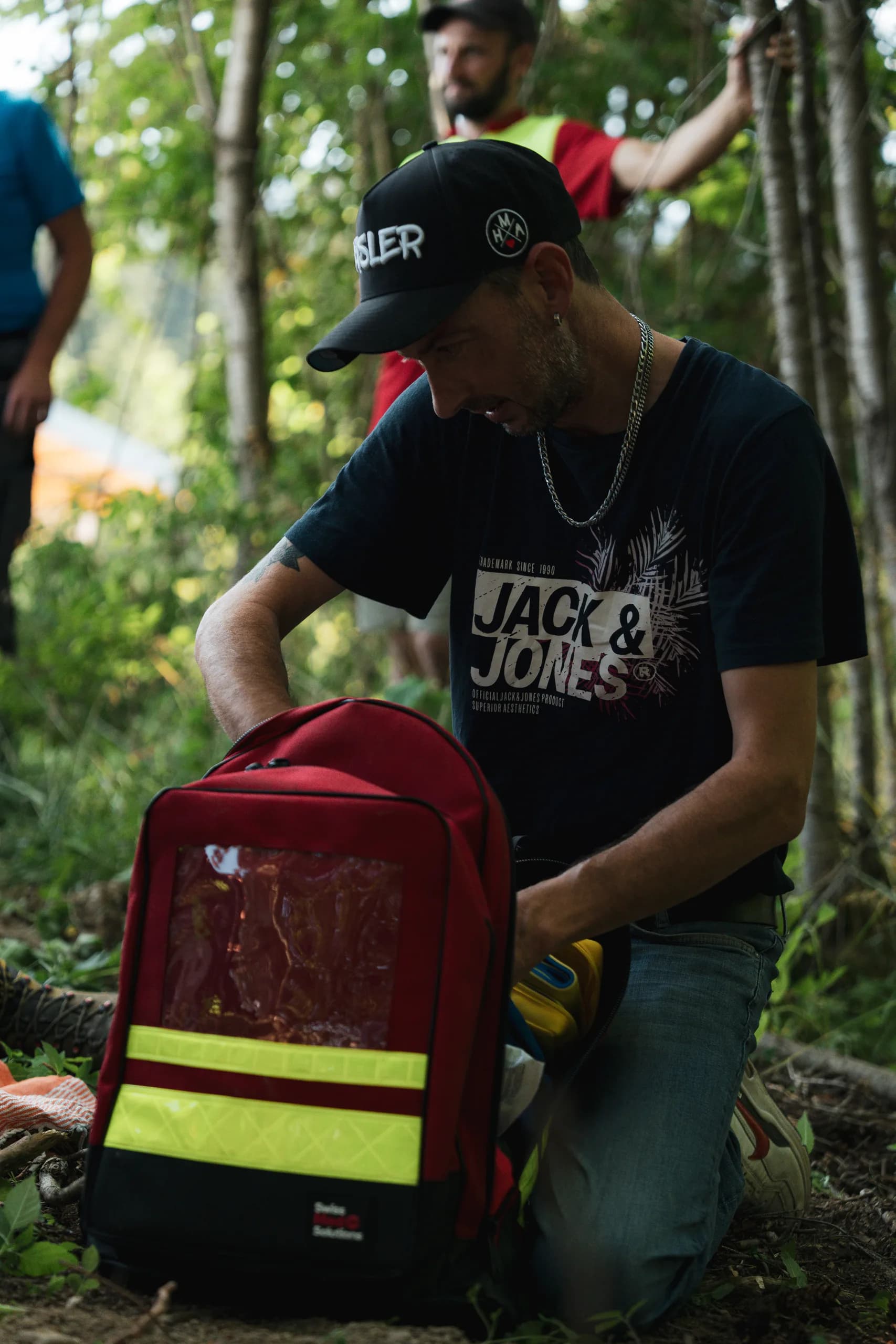 Un participant agenouillé en forêt ouvre un sac à dos de secours rouge à bandes réfléchissantes